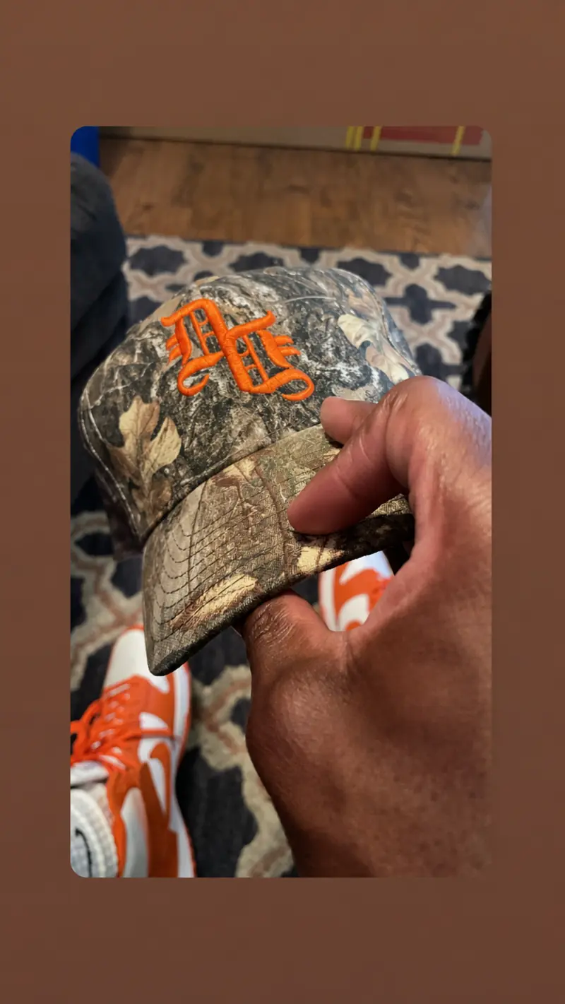 Daily Denim & Co. camouflage cap with orange embroidered logo held by the brim