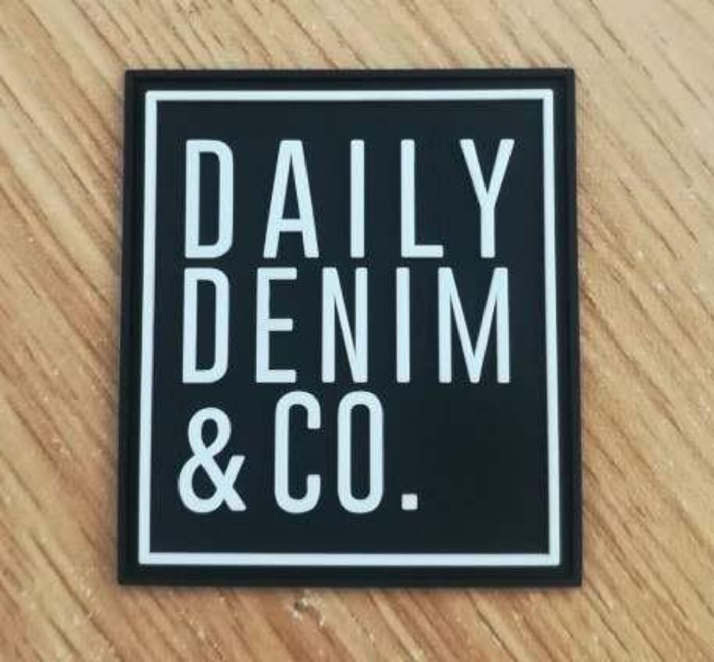 Physical black rubber Daily Denim & Co. label patch resting on a wooden surface